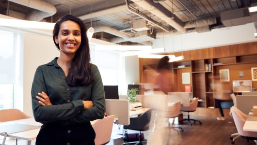 Portrait Of Smiling Businesswoman Standing In Busy Modern Open Plan Office