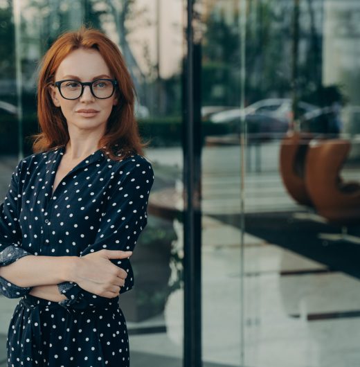 Self assured female director with red hair pleasant appearance dressed in elegant dress spectacles poses near office building waits for partner going to have meeting. Corporate worker outdoor