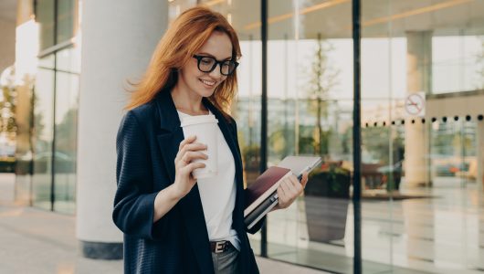 Smiling redhead woman corporate worker dressed in formal clothes drinks coffee carries notebook for making notes laptop focused down with happy expression poses near entrance of office building